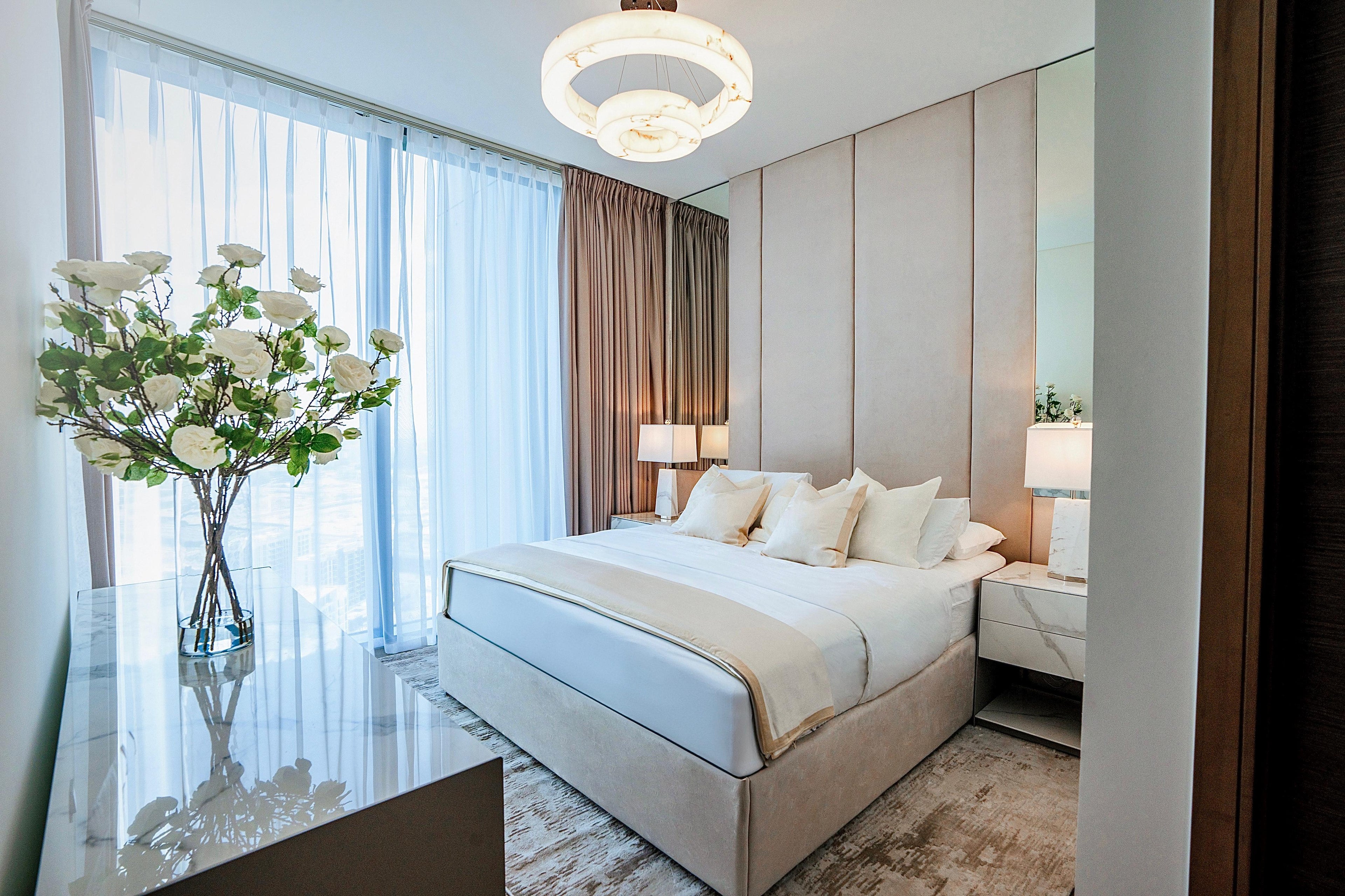 Elegant contemporary bedroom with a neutral upholstered bed, chandelier, and sheer white curtains.