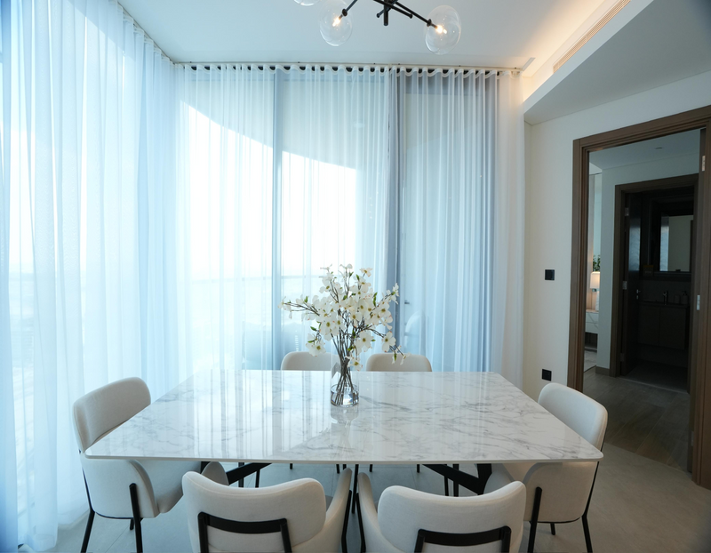 Modern apartment dining room interior design featuring a white marble table, soft curved chairs, sheer floor-to-ceiling curtains, and natural light.